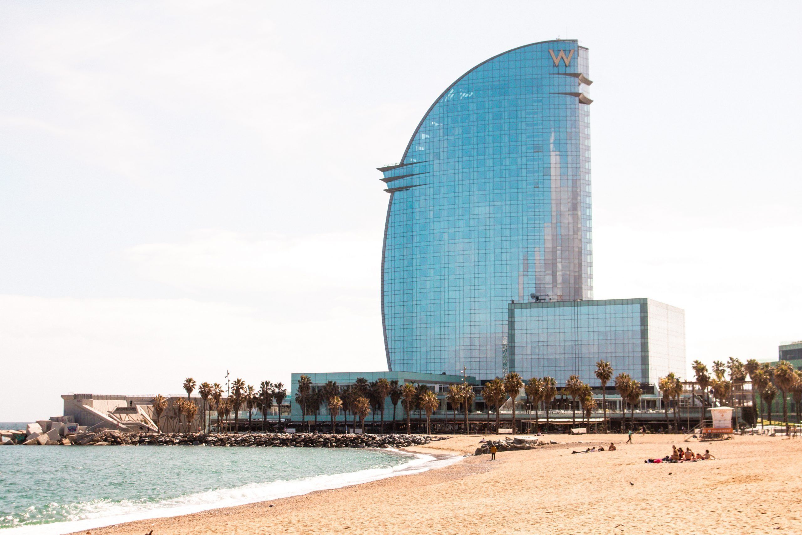An image of Barceloneta Beach in Barcelona featuring the iconic W Hotel and palm tree-lined promenade. 